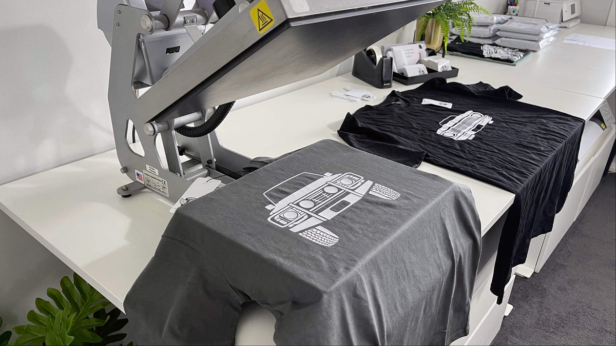 A gray t-shirt with a white graphic print is being held under a large heat press machine on a white worktable, with a black t-shirt featuring a similar graphic laid out nearby.