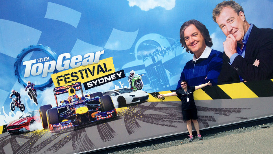 A large outdoor billboard or mural for the BBC Top Gear Festival Sydney, featuring racing cars, motorcycles, and a blue sky graphic, with two men standing in front of it outdoors.